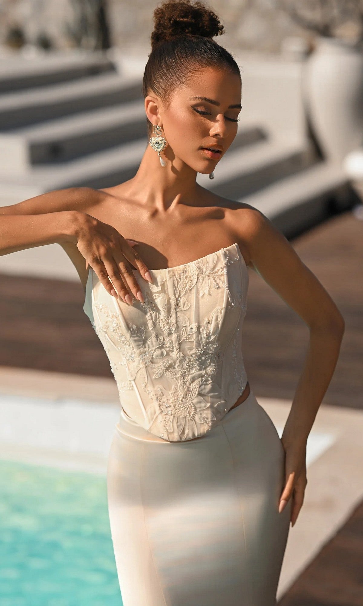 A woman in the Portia and Scarlett PS26092 long formal dress, featuring a strapless embroidered corset bodice, stands by a pool with one arm raised, wearing bold statement earrings and her hair styled in a high bun.